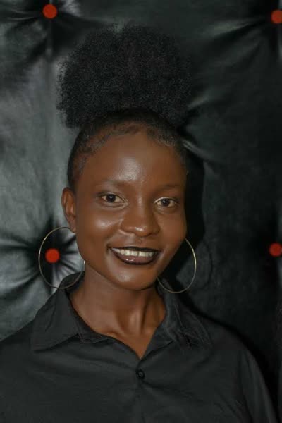 Studio headshot, smiling, hair worn up, large hoop earrings, against a dark padded backdrop. Lake City Studio, Kisumu 2023.